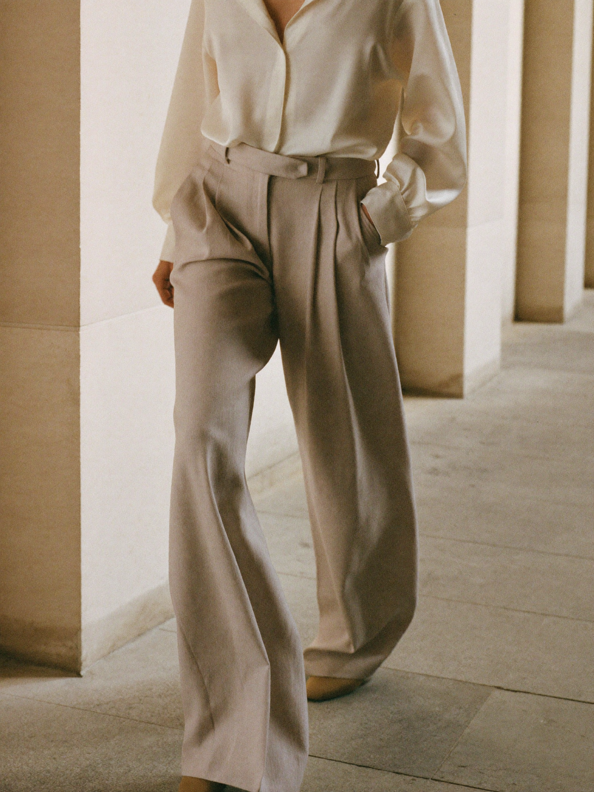 Wide-leg taupe linen trousers in motion along a sunlit colonnade